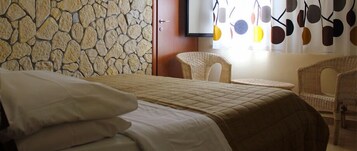 Un dormitorio con pared de piedra, cama con cubrecama acolchado, dos sillas de mimbre y una ventana con cortina de motivos circulares.