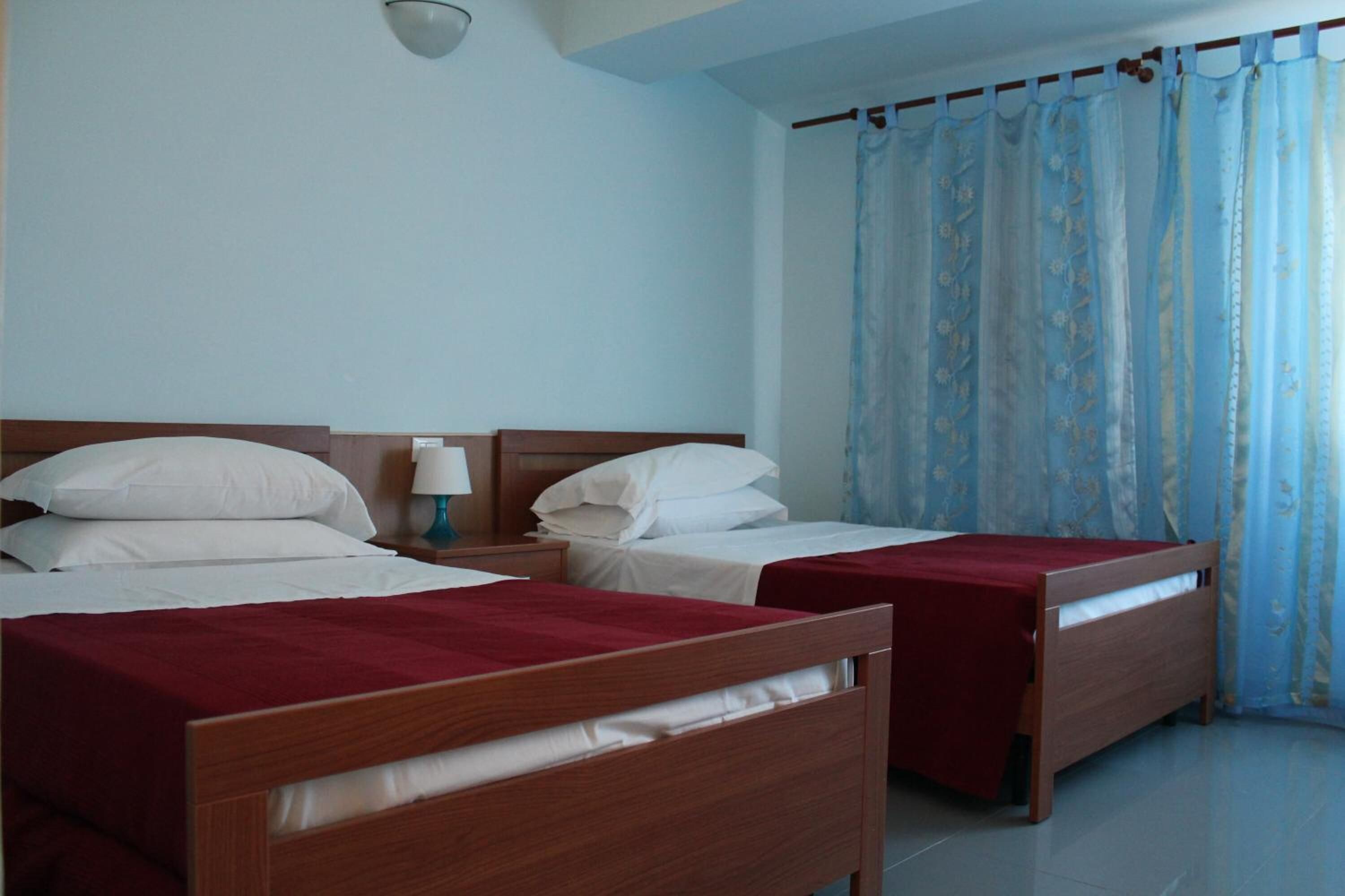 Una camera d'albergo con due letti in legno, biancheria da letto bianca, un comodino con una lampada e una finestra con tende blu.