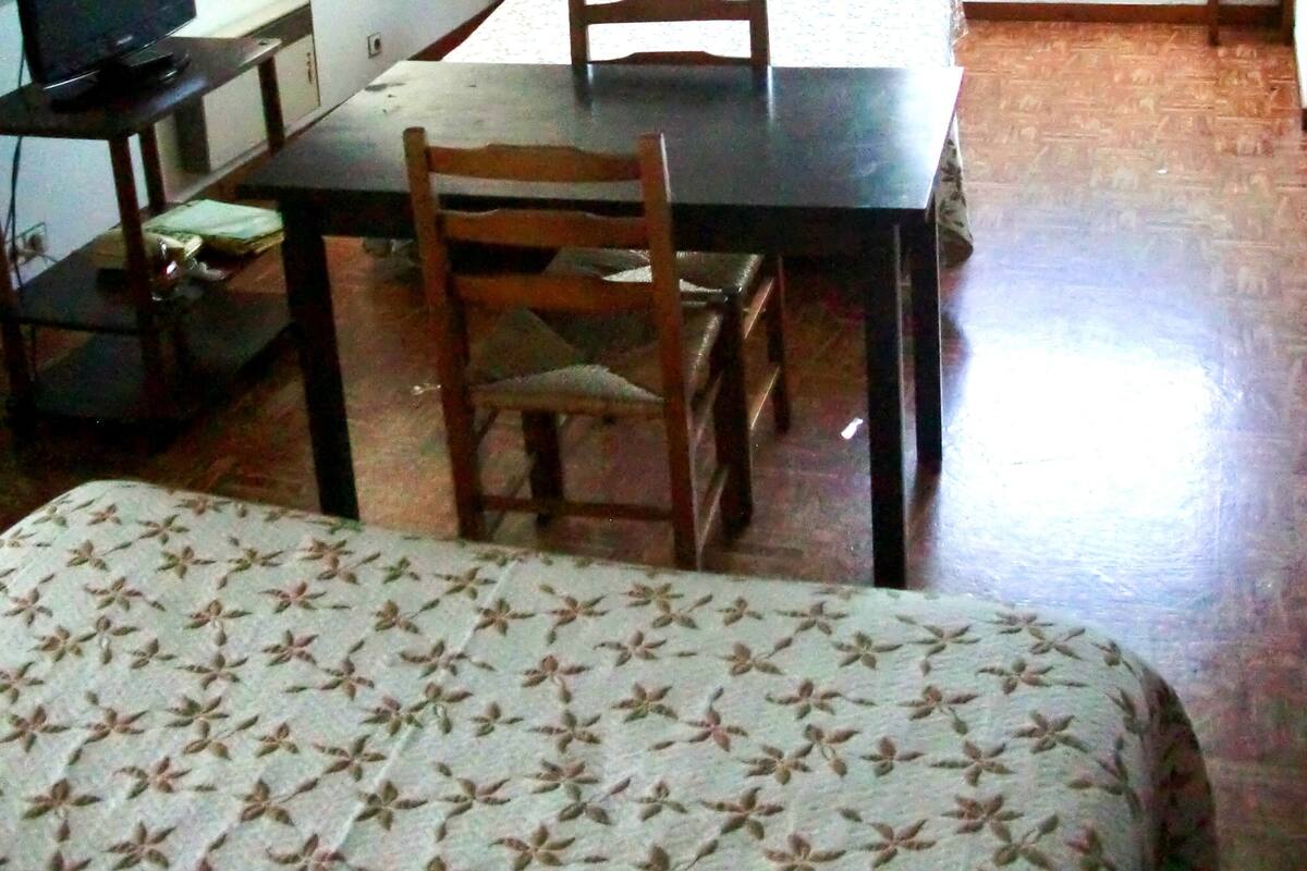 Ein kleiner, schwach beleuchteter Raum mit schräg stehender Decke, einem Bett, einem Schreibtisch mit Computer und einem Fenster, das natürliches Licht hereinlässt.