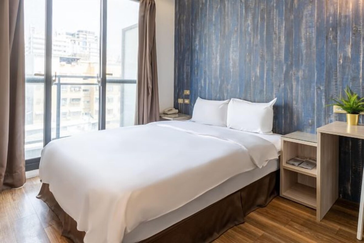 A hotel room with a large bed, a nightstand, a desk, and a view of the city through large windows.