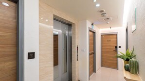 Área de elevadores moderna com duas elevadoras, uma porta de madeira e um pequeno vaso de planta numa prateleira.