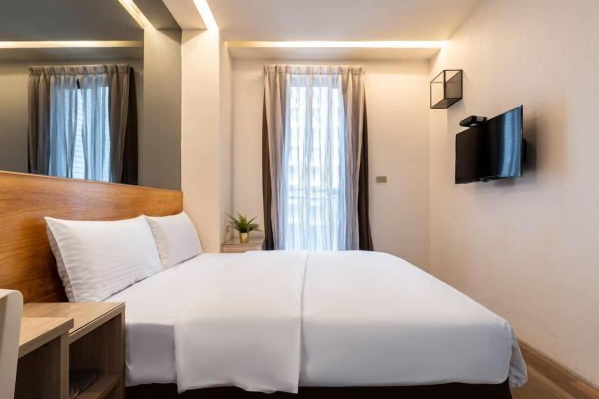 A hotel room with a large bed, a wooden headboard, a TV mounted on the wall, and a window with curtains.