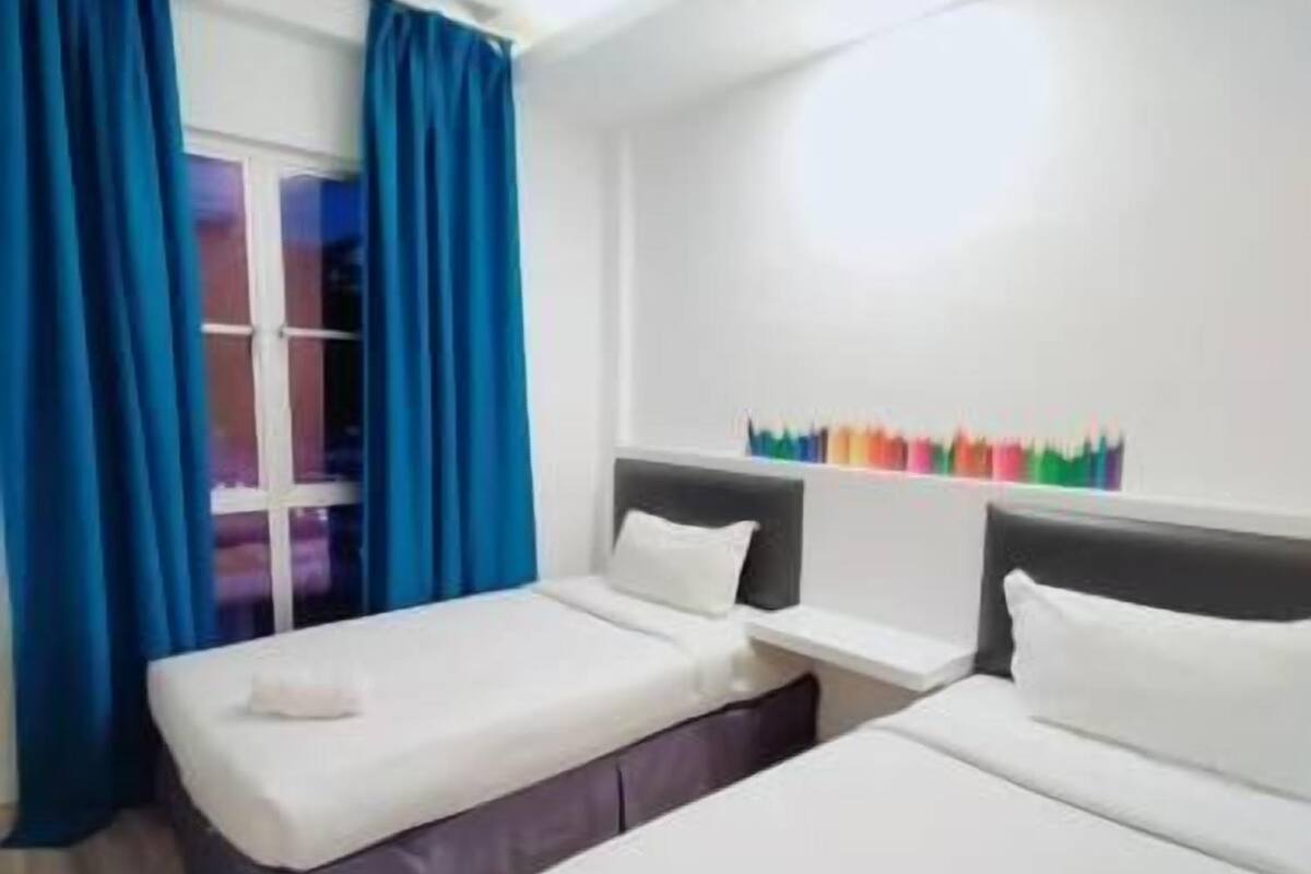 Ein Hotelzimmer mit zwei Betten, einem Fenster mit blauen Vorhängen, einem Nachttisch und einem Wandregal mit bunten Gegenständen.