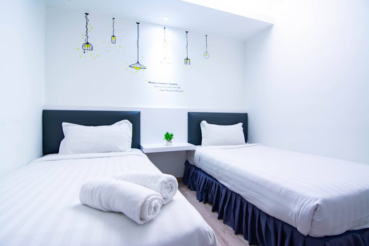 Ein Hotelzimmer mit zwei Betten, weißer Bettwäsche, einem kleinen Fenster mit Pflanze sowie einer Wand mit Leuchten und einem motivierenden Zitat.