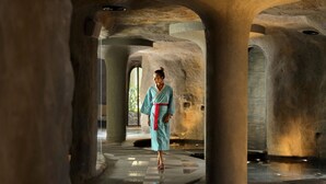 A person in traditional attire walking through a corridor with stone columns and a reflective floor. - Padma Resort Ubud (Payangan)