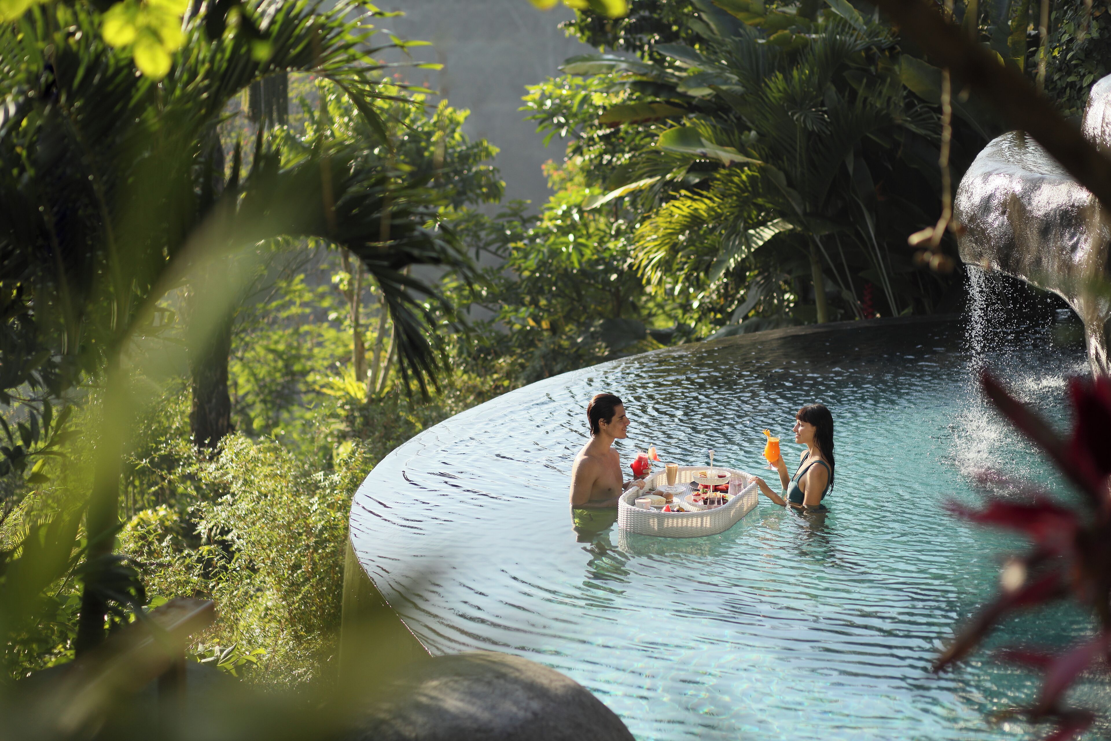 Una pareja en una piscina circular rodeada de exuberante vegetación y una gran formación rocosa.