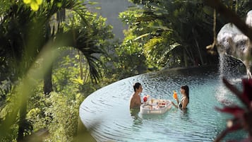 Una pareja en una piscina circular rodeada de exuberante vegetación y una gran formación rocosa.