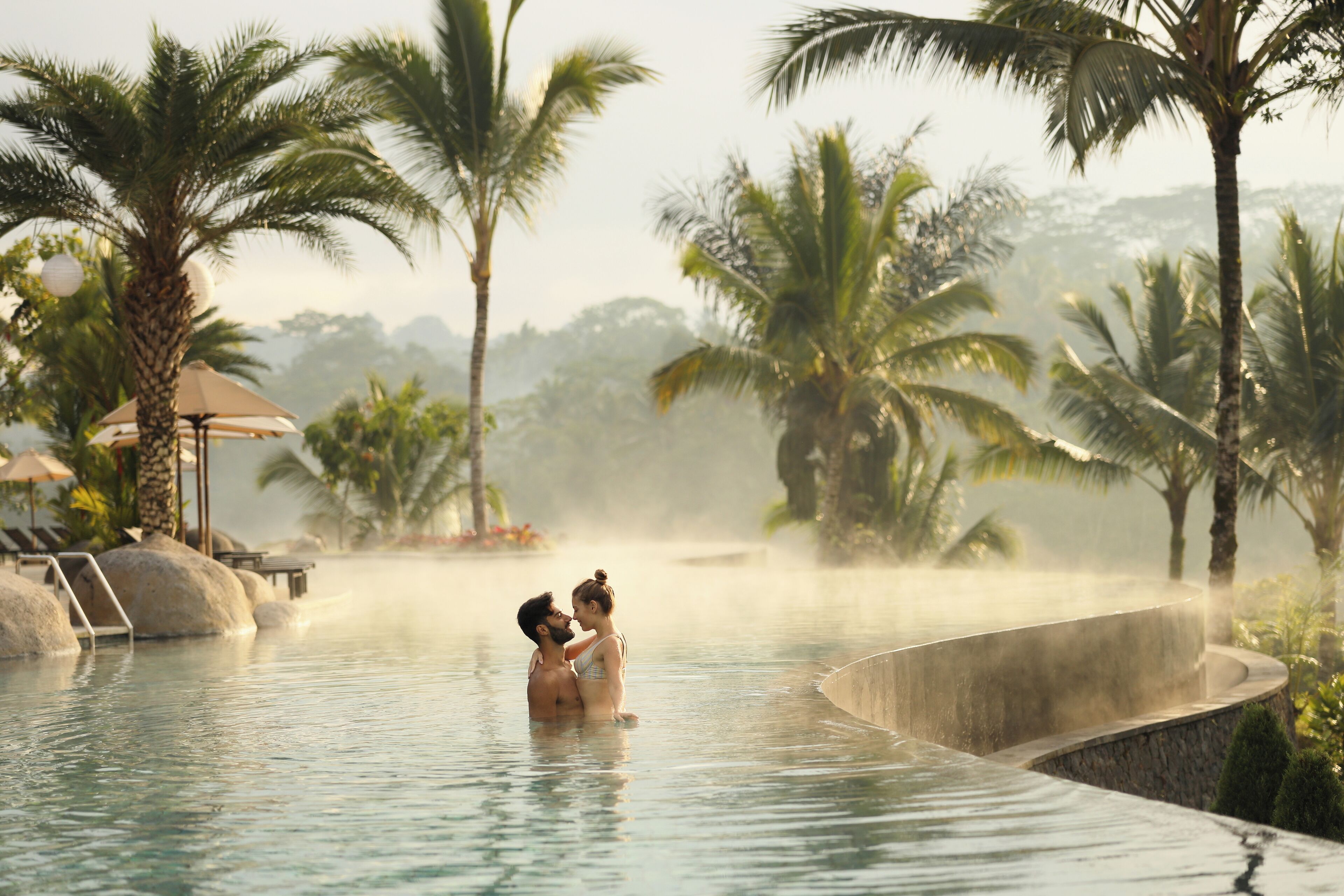 A couple in a heated pool surrounded by palm trees and lounge chairs.