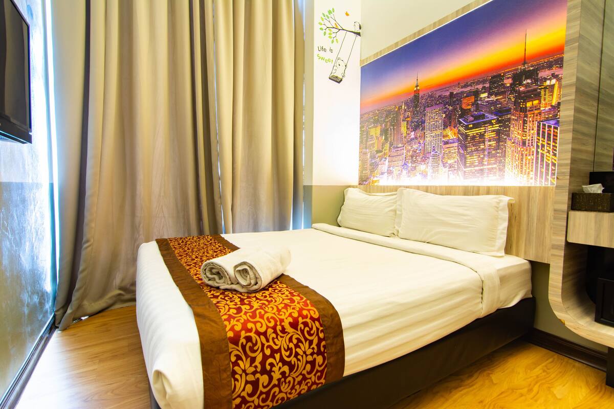 A neatly made bed with a patterned bedspread, a window with curtains, and a cityscape mural on the wall.
