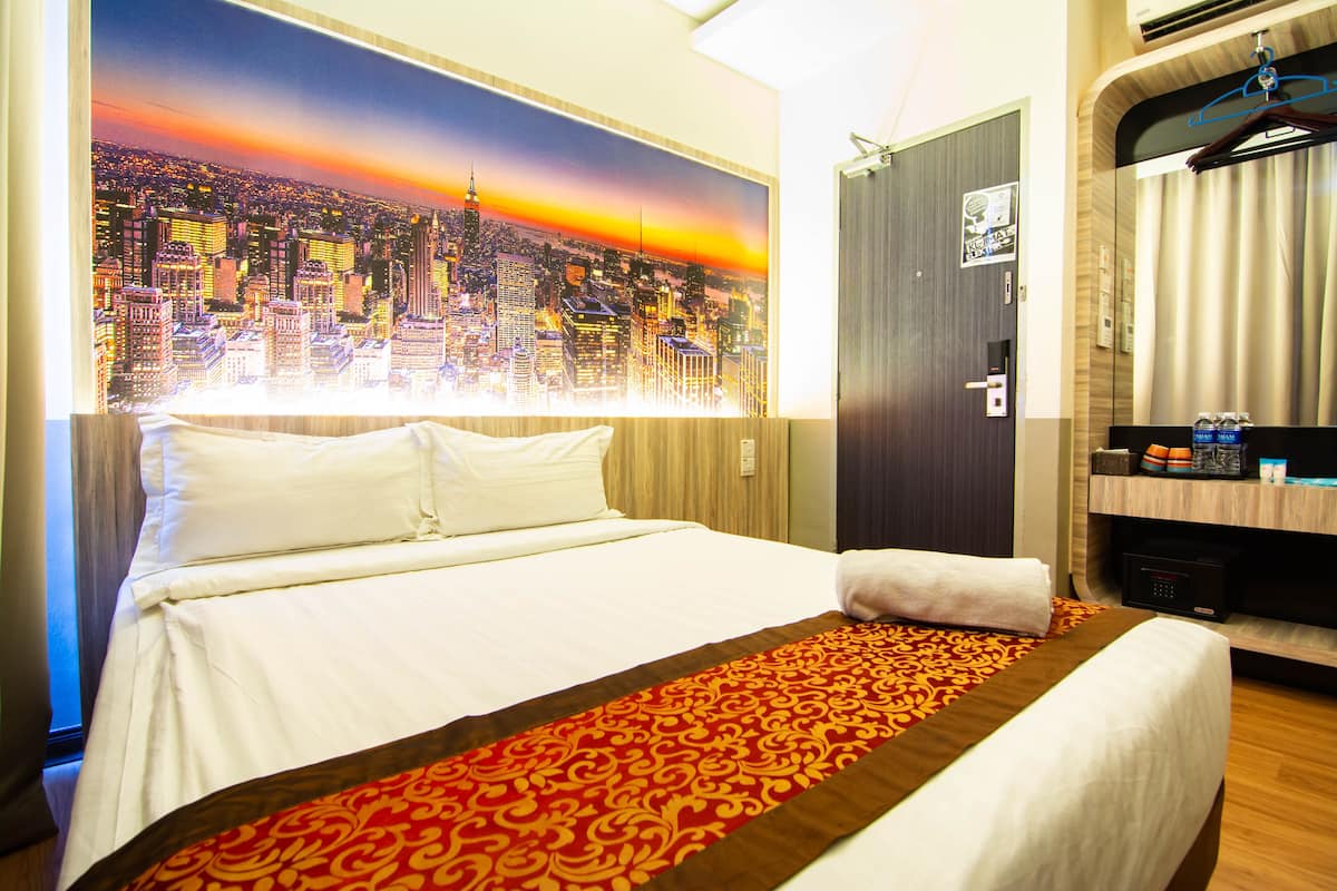 A hotel room with a large bed, a cityscape painting on the wall, a wooden headboard, a mirror, and a shelf with toiletries.