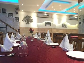 Un comedor con mesas dispuestas para una comida, con cubertería de vidrio transparente, servilletas blancas y mantel rojo.