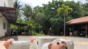 A courtyard with large, textured sheep sculptures, surrounded by lush greenery and a building with a red roof. - Papangkorn House (Surat Thani)