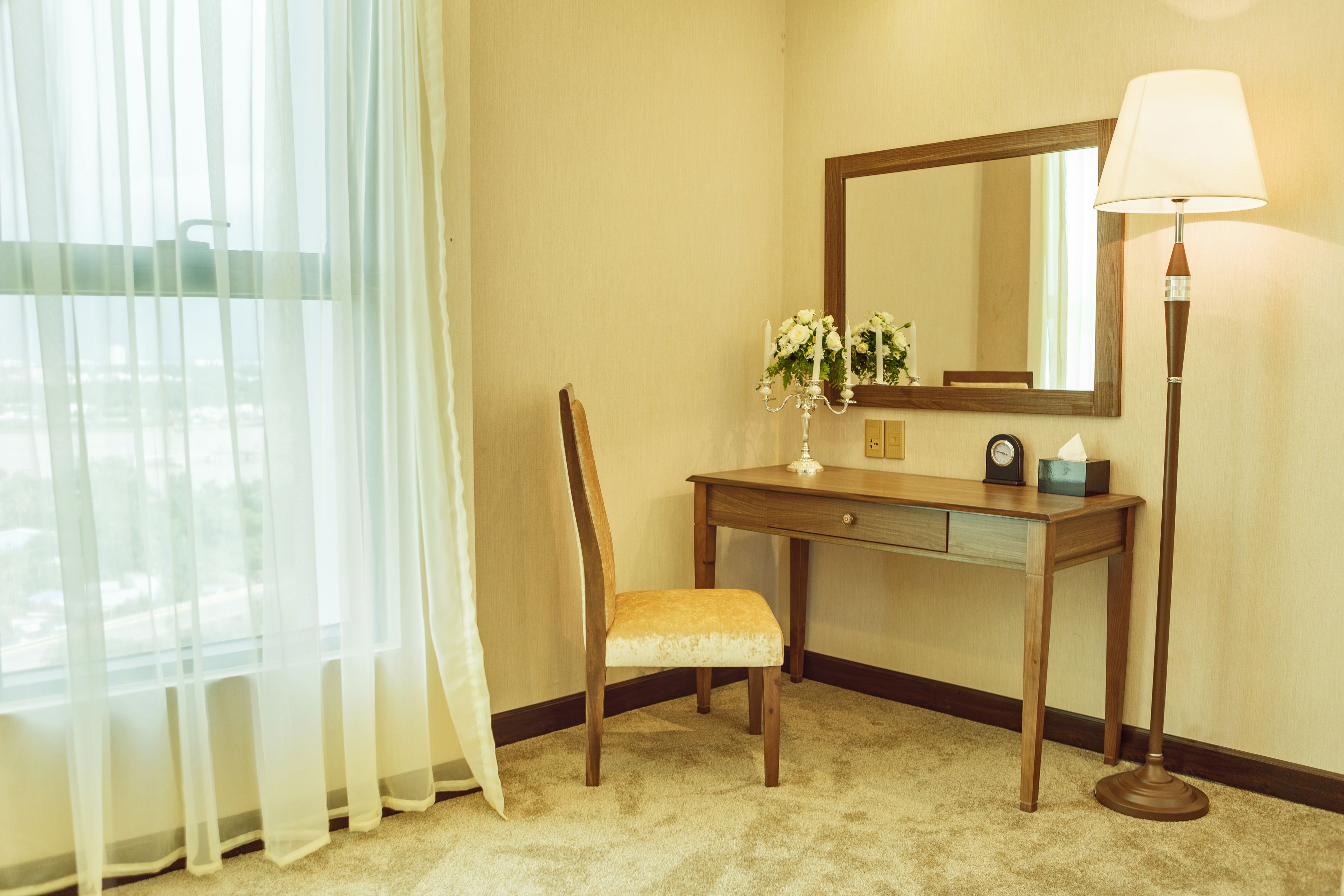 A hotel room with a wooden desk, a chair, a mirror, a lamp, and a window with sheer curtains.