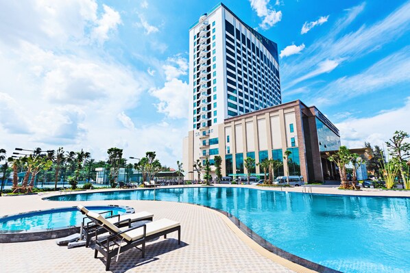 Un hotel con una piscina grande, sillas de descanso, palmeras y un edificio alto al fondo.