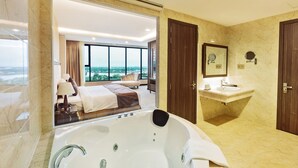 Ein modernes Badezimmer mit einer großen Badewanne, einem Waschtisch inklusive Spiegel und Blick auf ein Schlafzimmer mit Bett und Fenster.