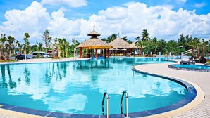 Ein großer Pool im Freien mit einem pavilons mit Reetdach, Palmen und blauem Himmel.