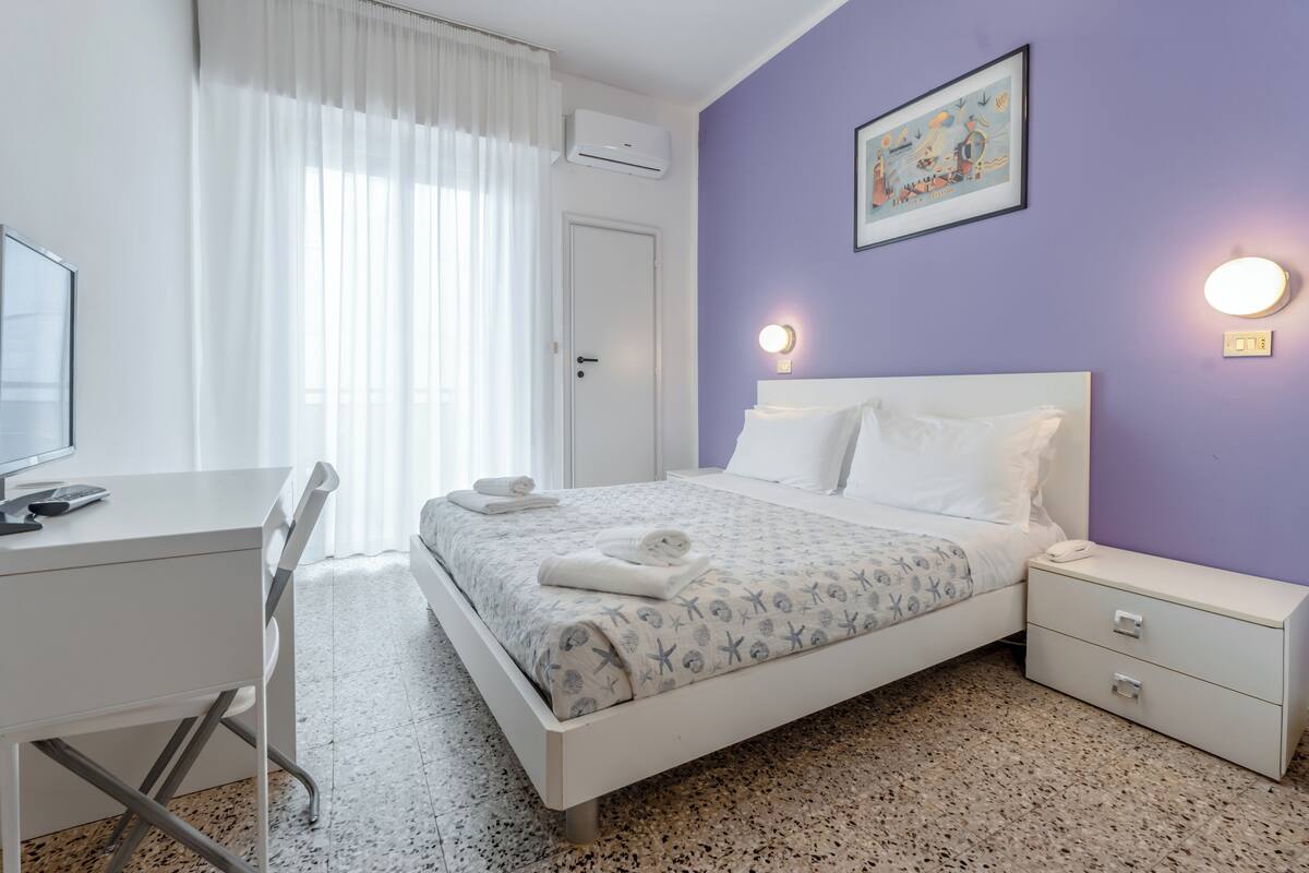 Un dormitorio con cama, mesita de noche, escritorio, silla y una ventana con cortinas.