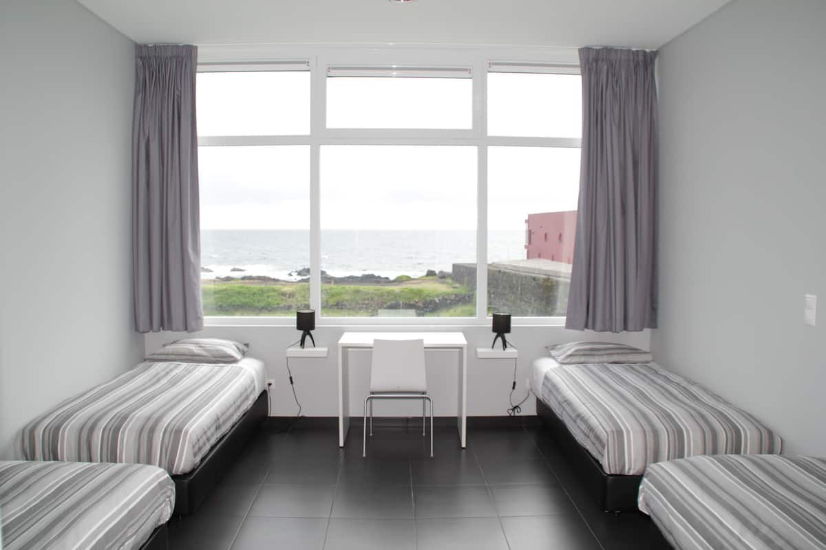Una habitación con dos camas, un escritorio y una silla, con vista al mar y un edificio al fondo.