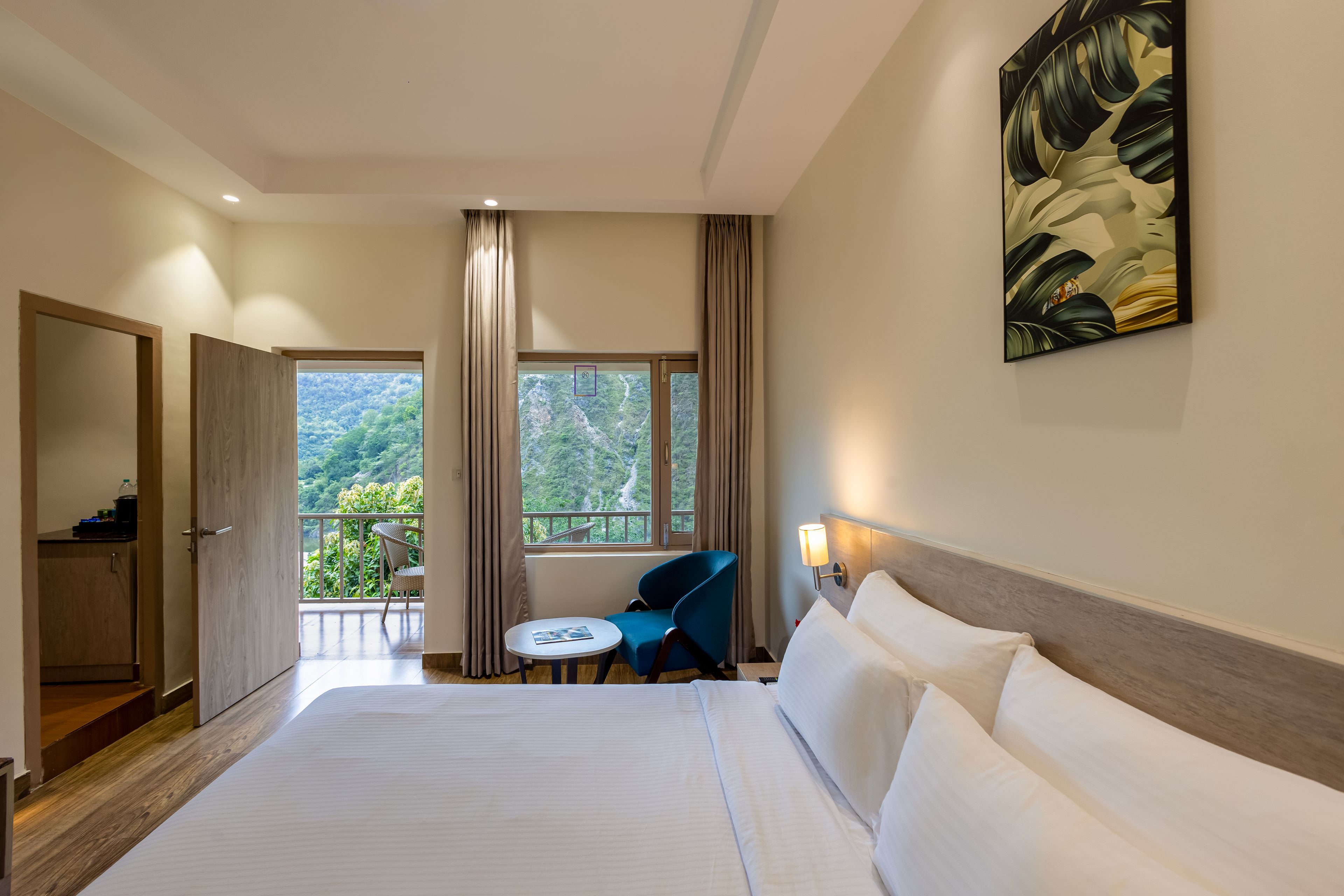 A hotel room with a large bed, a small table, a chair, a wardrobe, and a balcony with a view of greenery.