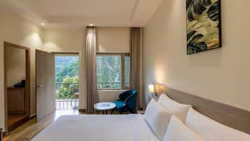 A hotel room with a large bed, a small table, a chair, a wardrobe, and a balcony with a view of greenery.