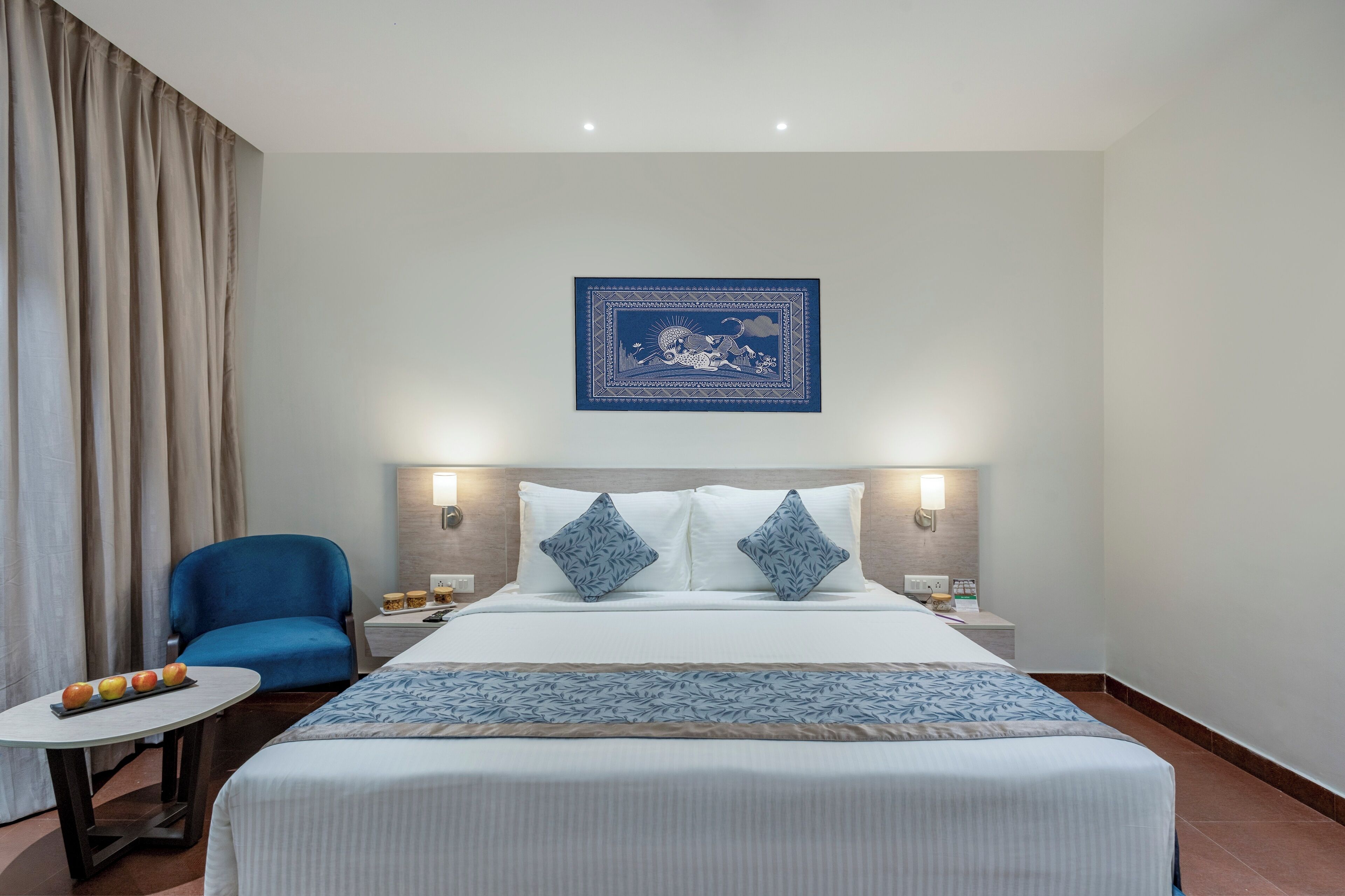 A hotel room with a bed, two pillows, a nightstand, a chair, a table with fruit, and a wall hanging.