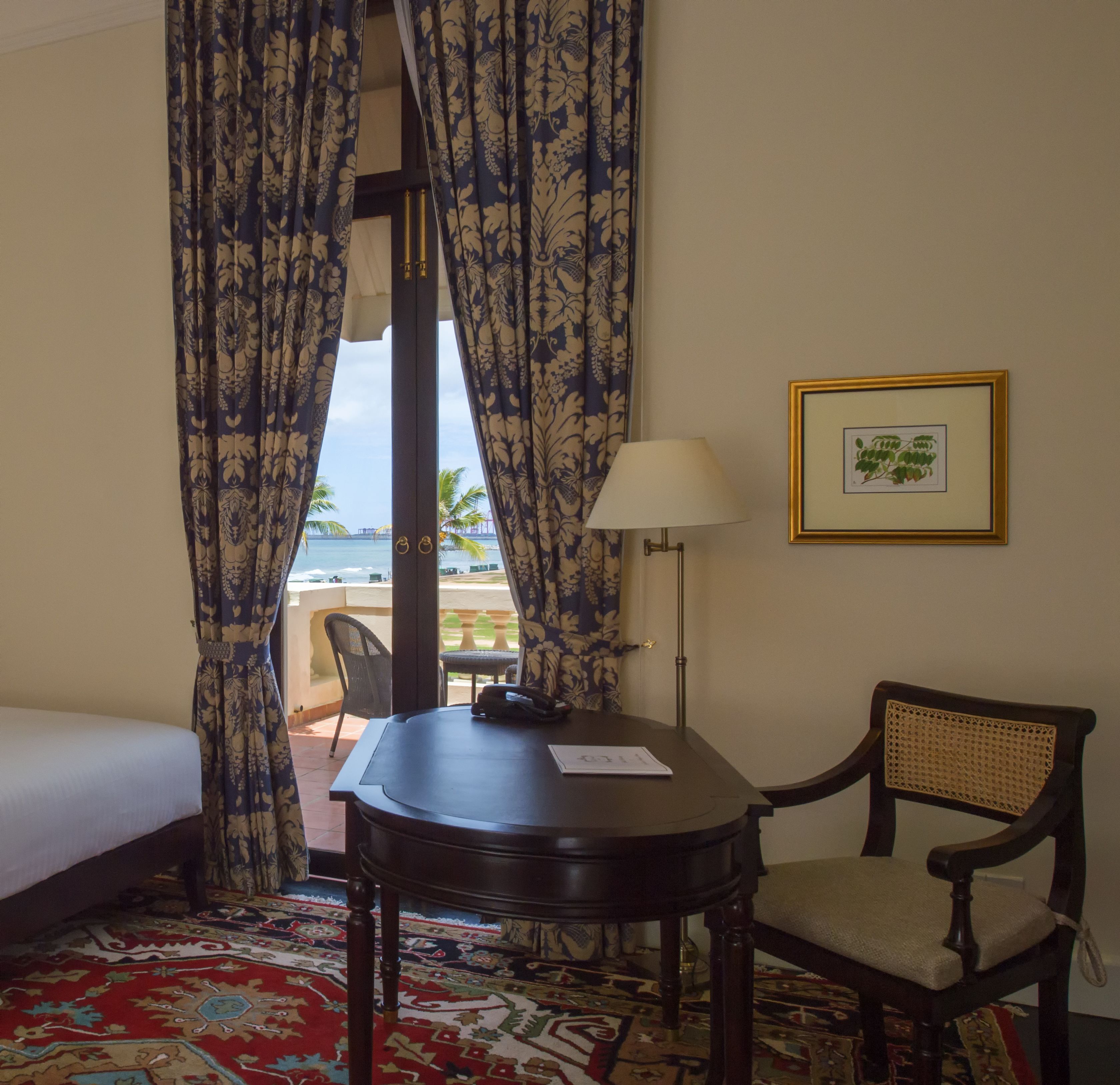 A hotel room with a bed, a table with a chair, a lamp, and a framed picture on the wall.