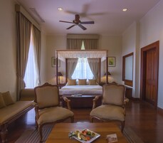 Habitación de hotel con cama con dosel, dos sillones, una mesa de centro de madera con fruta y un ventilador de techo.