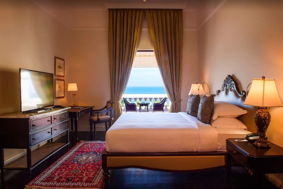 A hotel room with a large bed, a desk with a television, a chair, a nightstand with a lamp, and a view of the sea through a window with curtains.