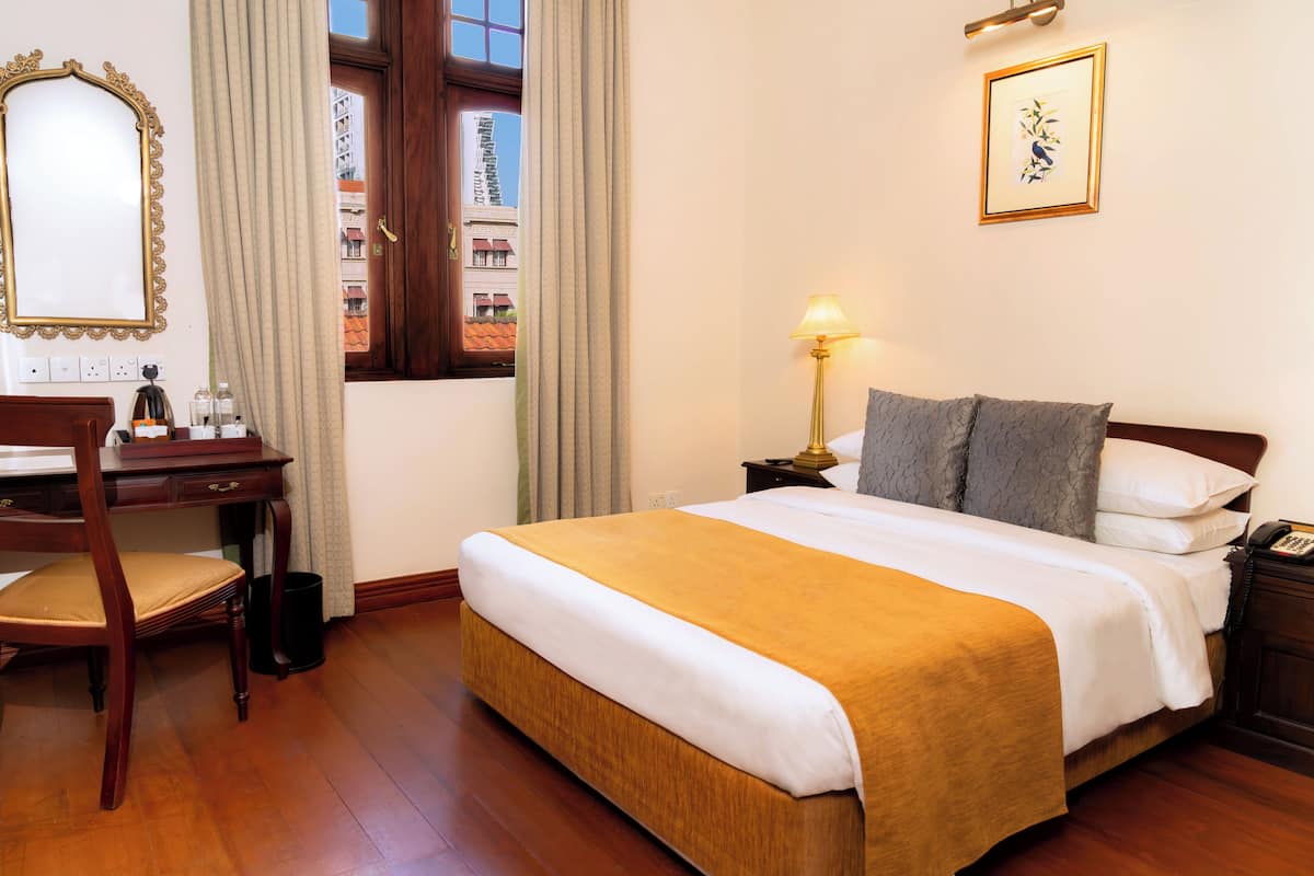 A hotel room with a large bed, a desk with a chair, a nightstand with a lamp, a framed picture on the wall, and a window with curtains.