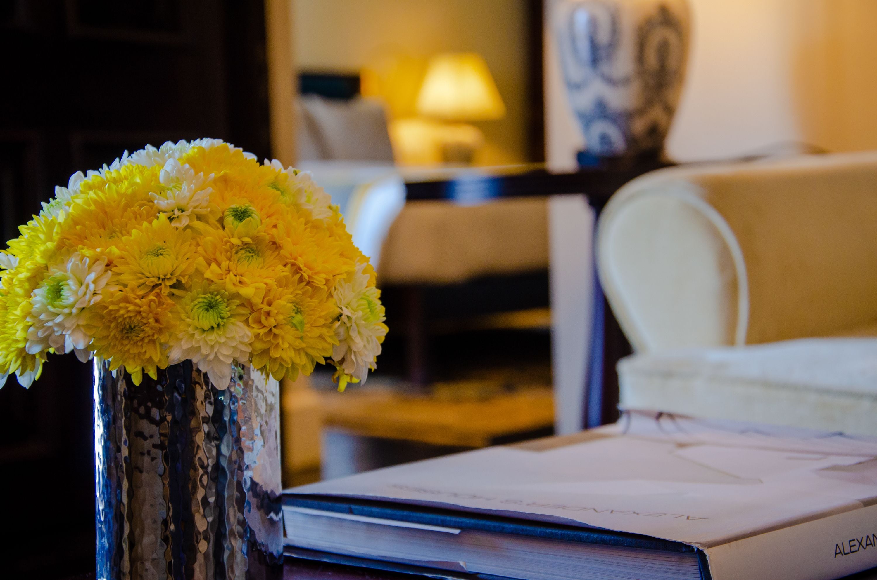 A room with a vase of yellow flowers on a table, a lamp, and a chair.