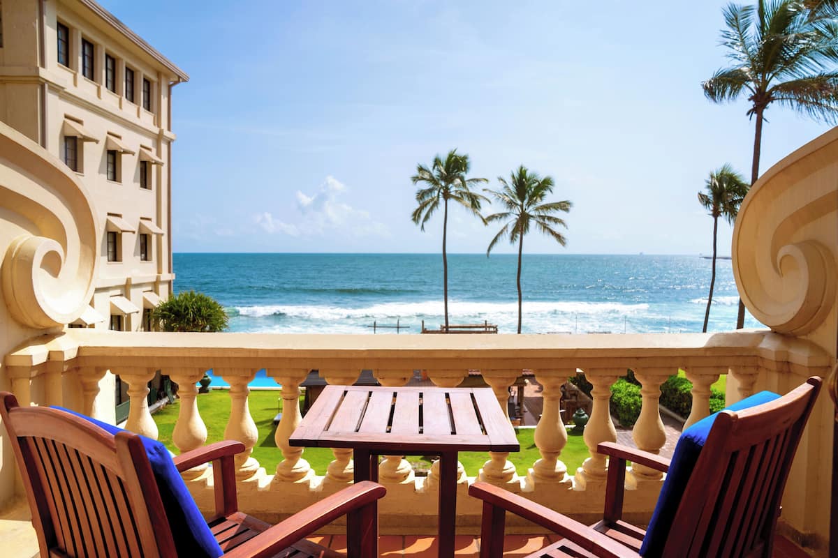 A balcony with a wooden table and chairs, overlooking the ocean with palm trees and a building in the background.