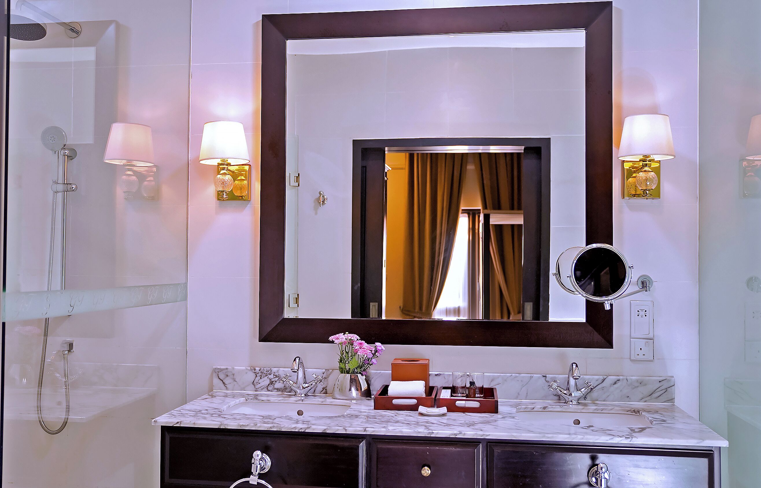 A bathroom with a marble sink, two wall-mounted lamps, a large mirror, and a shower area.