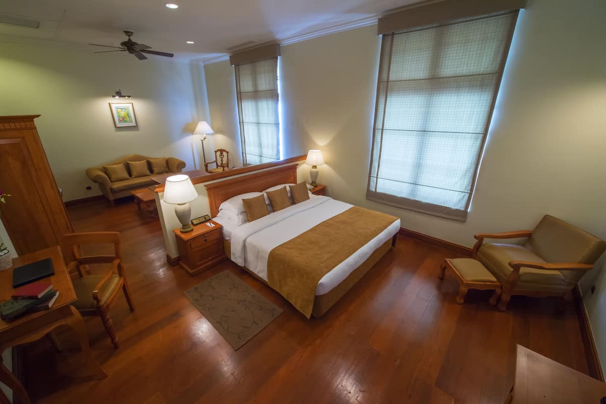 A hotel room with a large bed, a wooden desk, a chair, a sofa, and two windows with blinds.