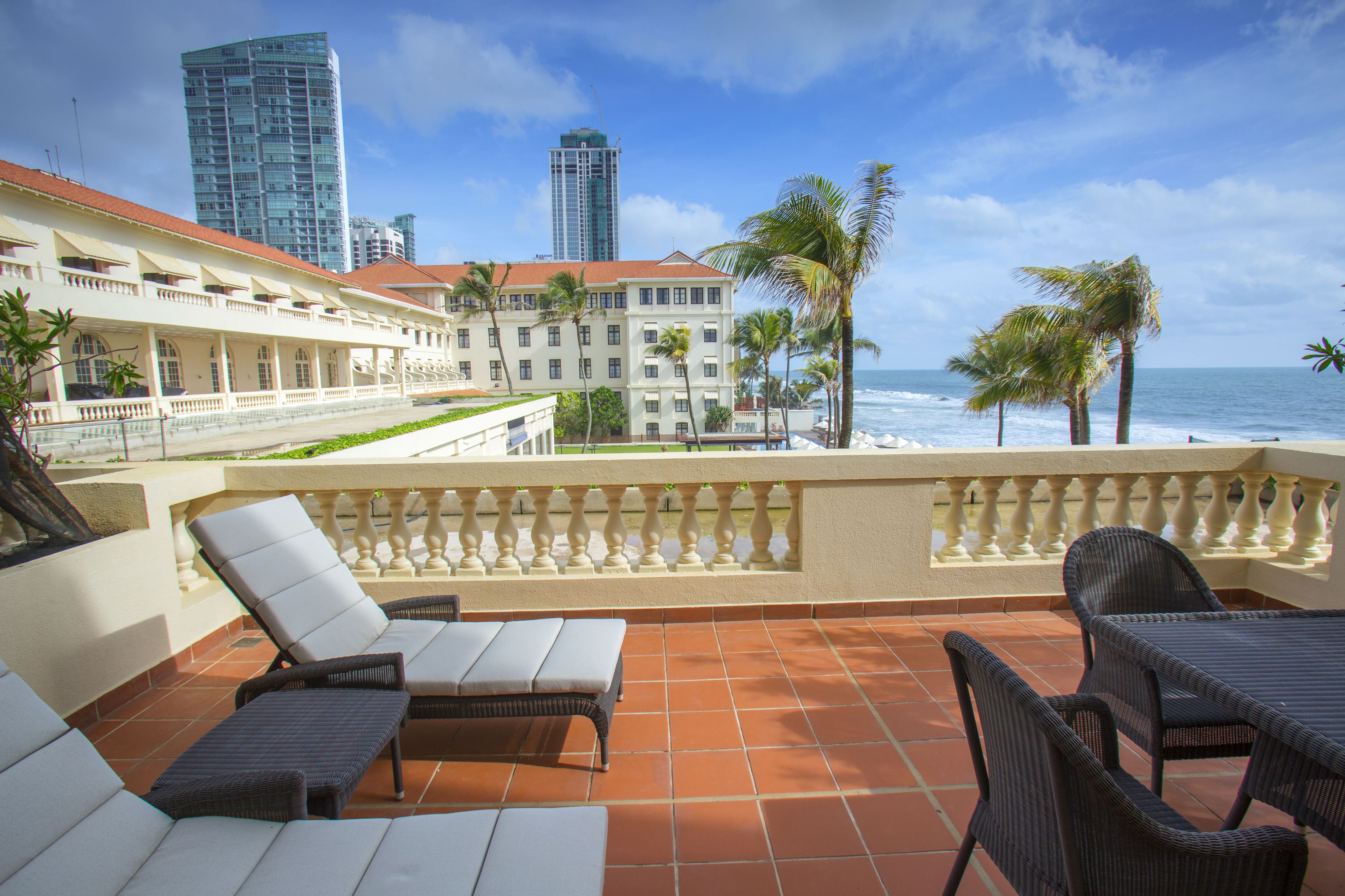 A terrace with lounge chairs and a table, overlooking a beach and palm trees, with a cityscape in the background.