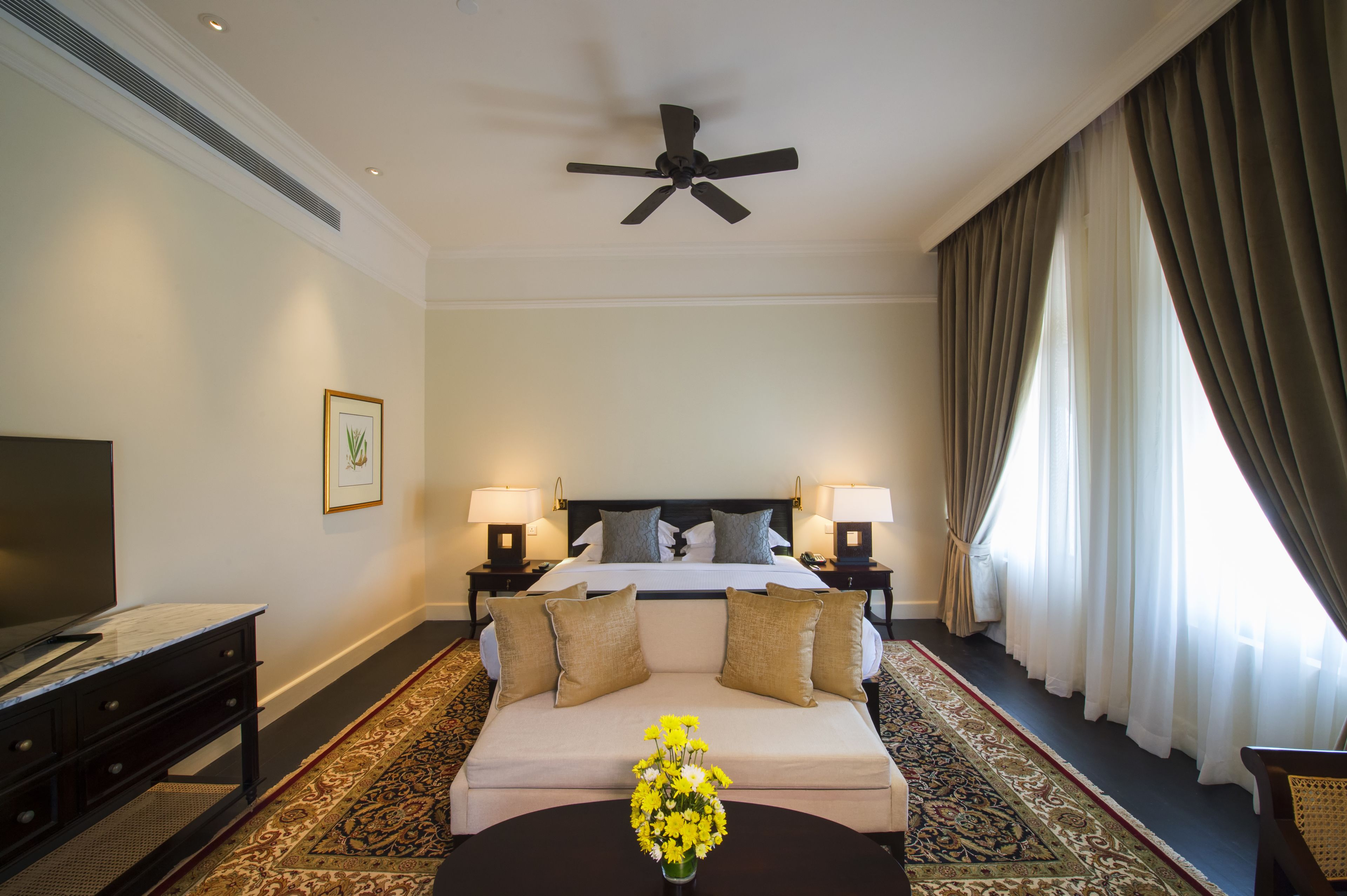 A hotel room with a bed, two bedside lamps, a ceiling fan, a TV, a sofa, a coffee table with flowers, and a large window with curtains.