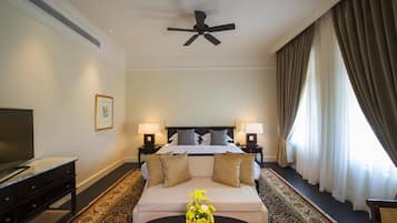 A hotel room with a bed, two bedside lamps, a ceiling fan, a TV, a sofa, a coffee table with flowers, and a large window with curtains.
