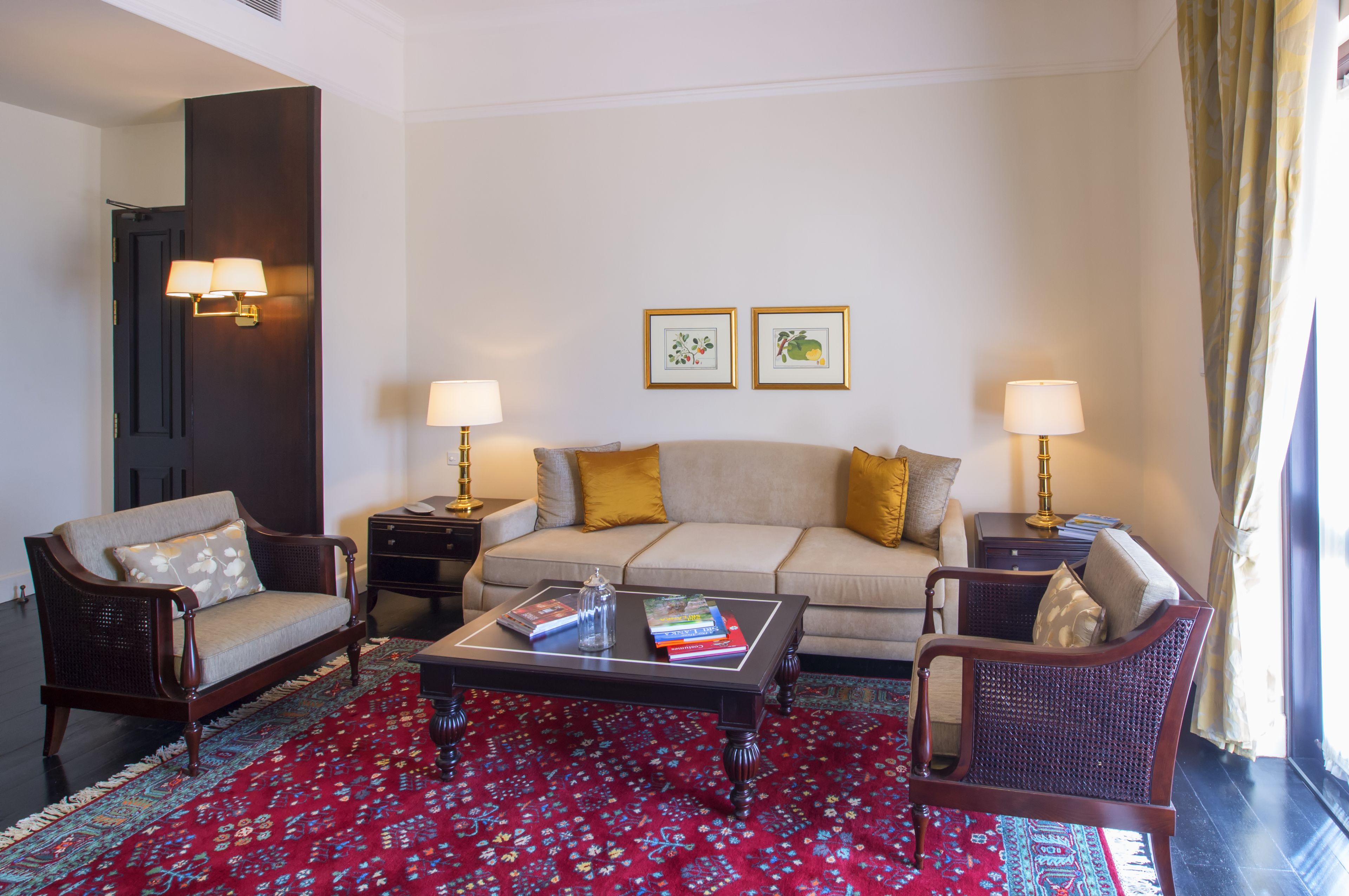 A living room with a sofa, two armchairs, a coffee table, and two side tables with lamps. There are framed pictures on the wall and a red patterned rug on the floor.