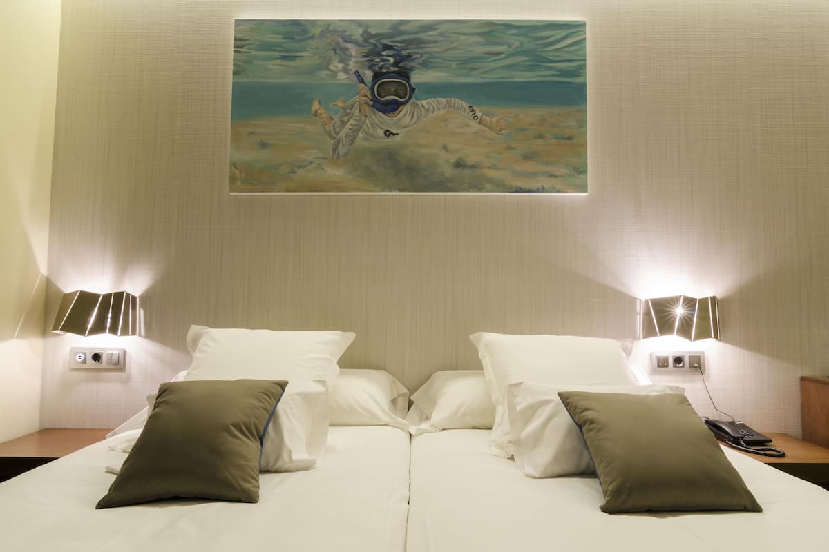 Una habitación de hotel con una cama, dos lámparas de noche, un cuadro de una escena submarina y una mesita de noche con un teléfono.