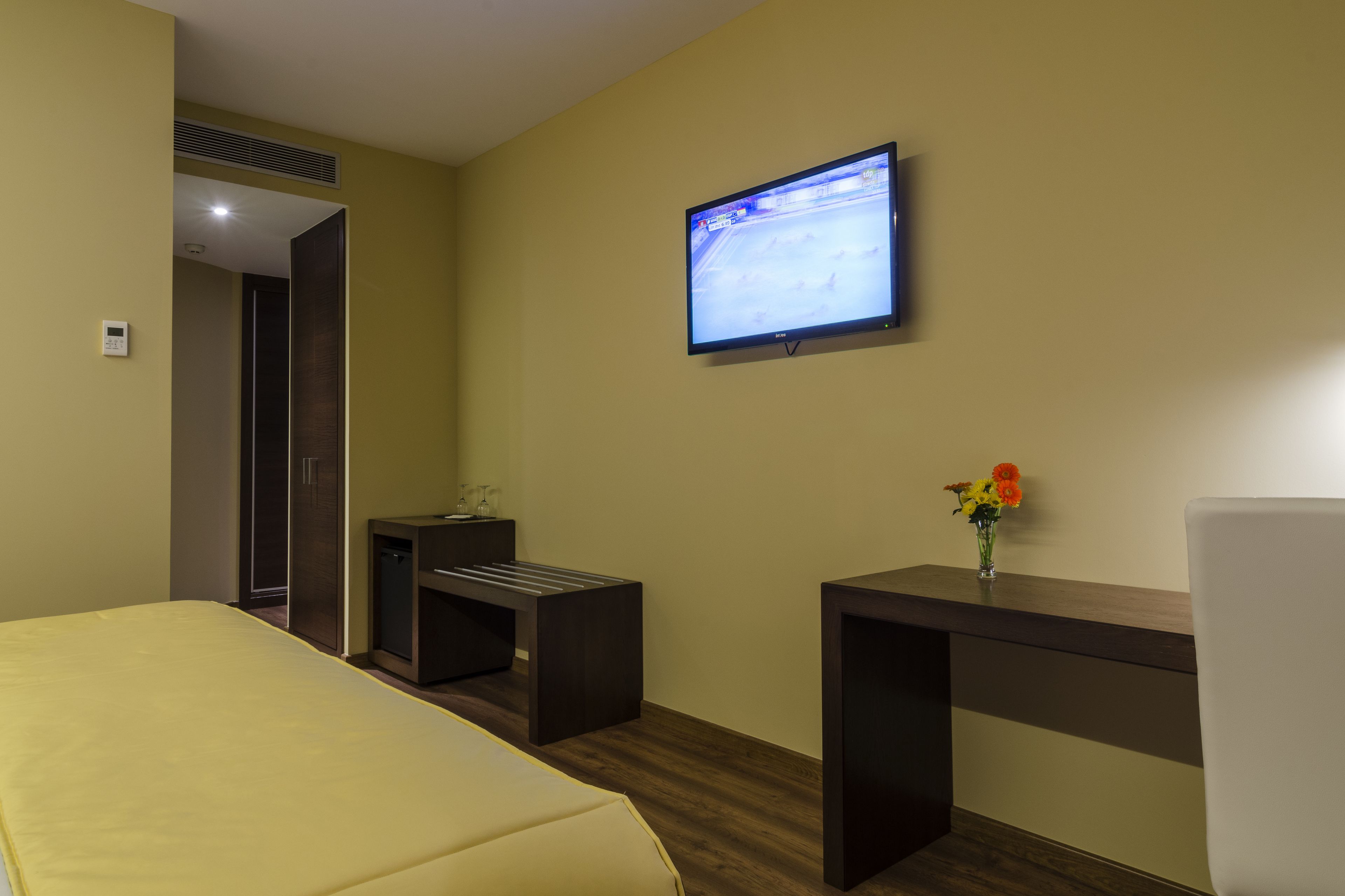A hotel room with a bed, a desk, a chair, a TV mounted on the wall, and a small table with a vase of flowers.