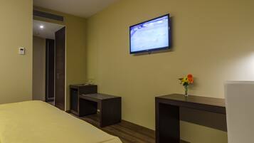 A hotel room with a bed, a desk, a chair, a TV mounted on the wall, and a small table with a vase of flowers.