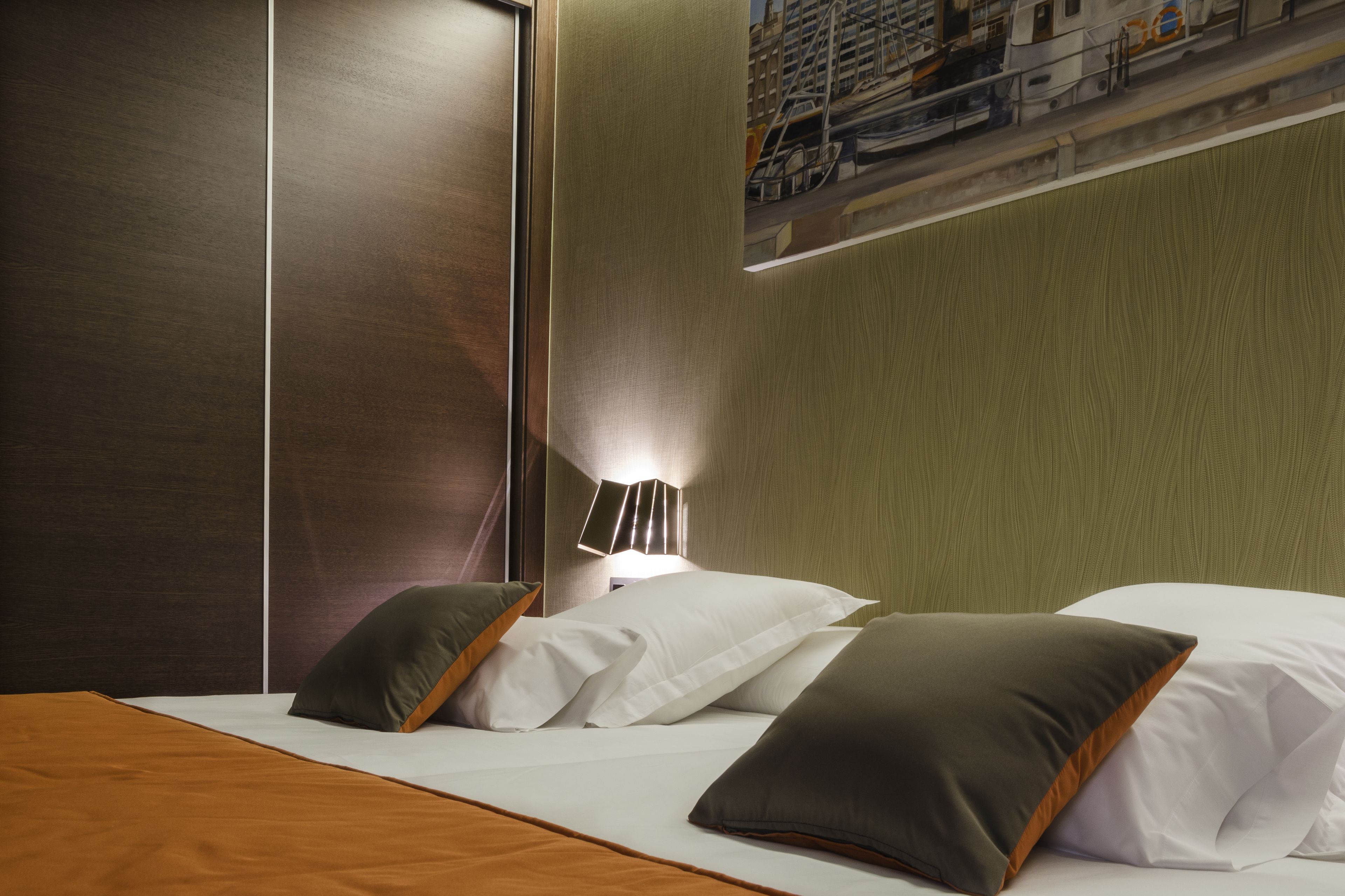 Habitación de hotel con una cama, almohadas, una mesita de noche, una lámpara y una obra de arte colgada en la pared.