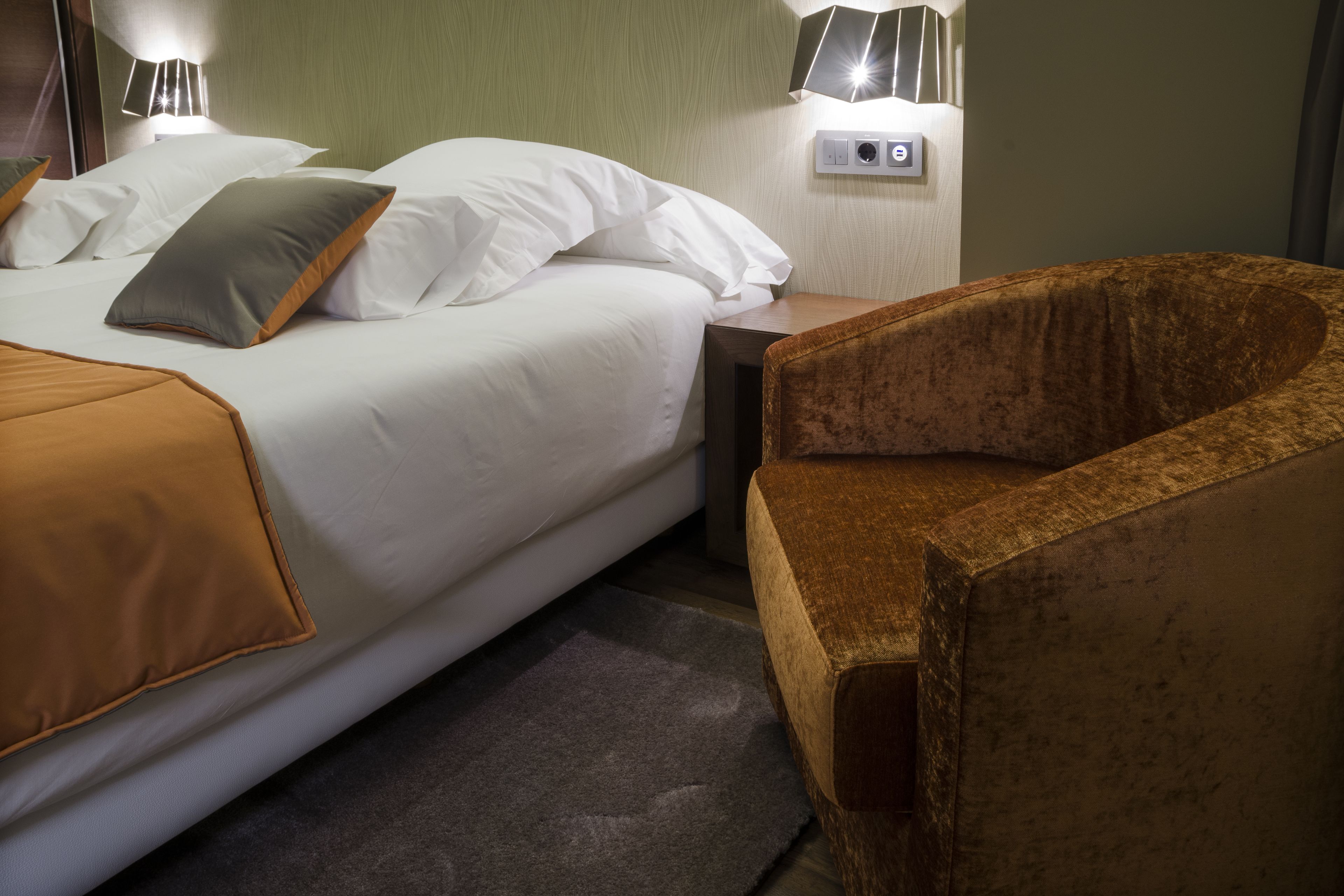 Una cama bien tendida con sábanas blancas y una manta marrón, un sillón tapizado en marrón y una mesita de noche con una lámpara y un enchufe.