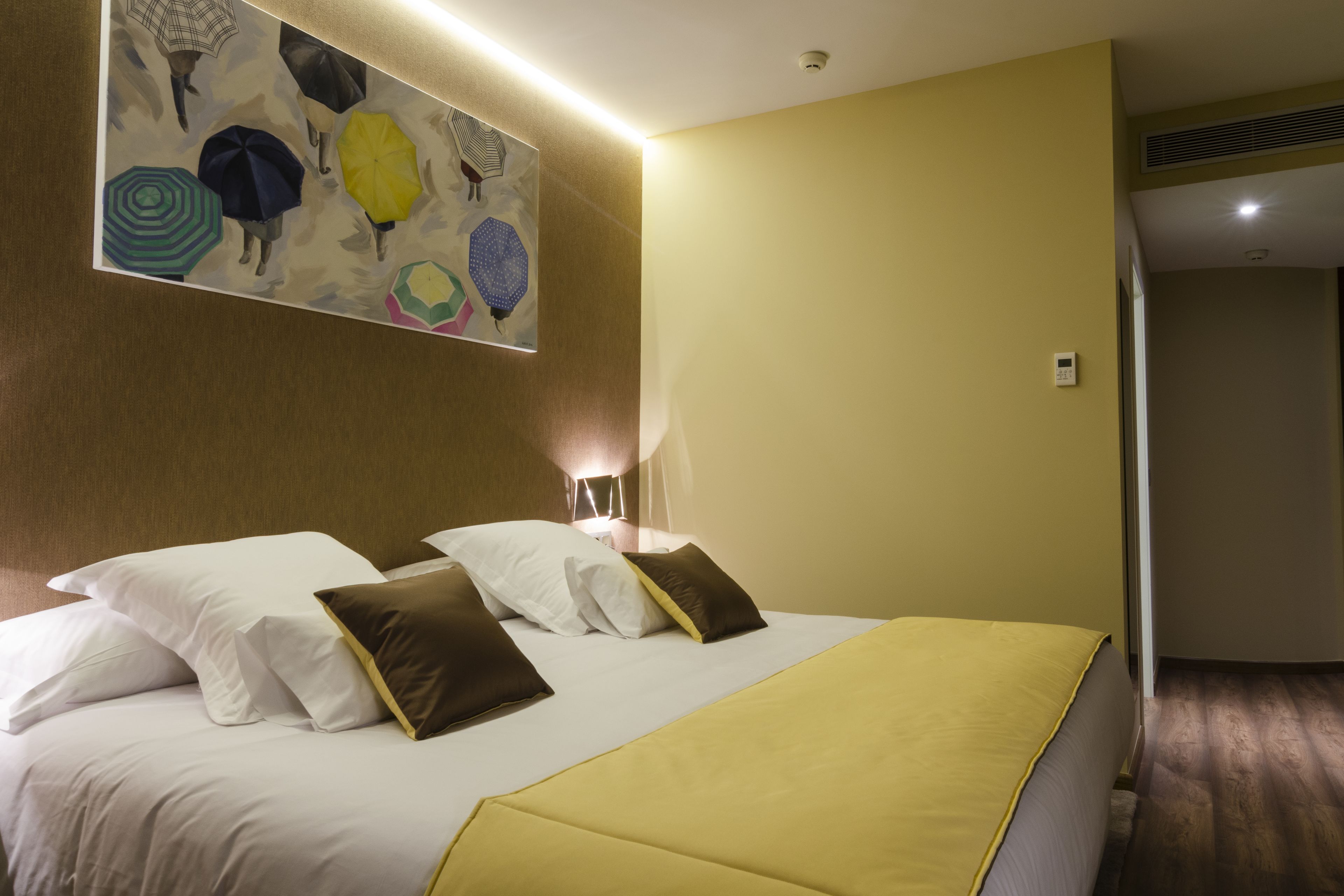 Quarto de hotel com uma cama grande, almofadas brancas e castanhas, colcha amarela e uma obra de arte na parede com guarda-chuvas.