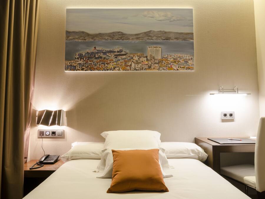 Una habitación de hotel con una cama, una mesita de noche con teléfono, una lámpara y un cuadro de una ciudad costera.