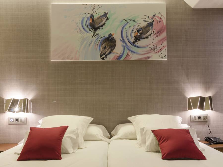 Quarto de hotel com uma cama, dois travesseiros vermelhos, um quadro pendurado na parede e abajures de cabeceira.