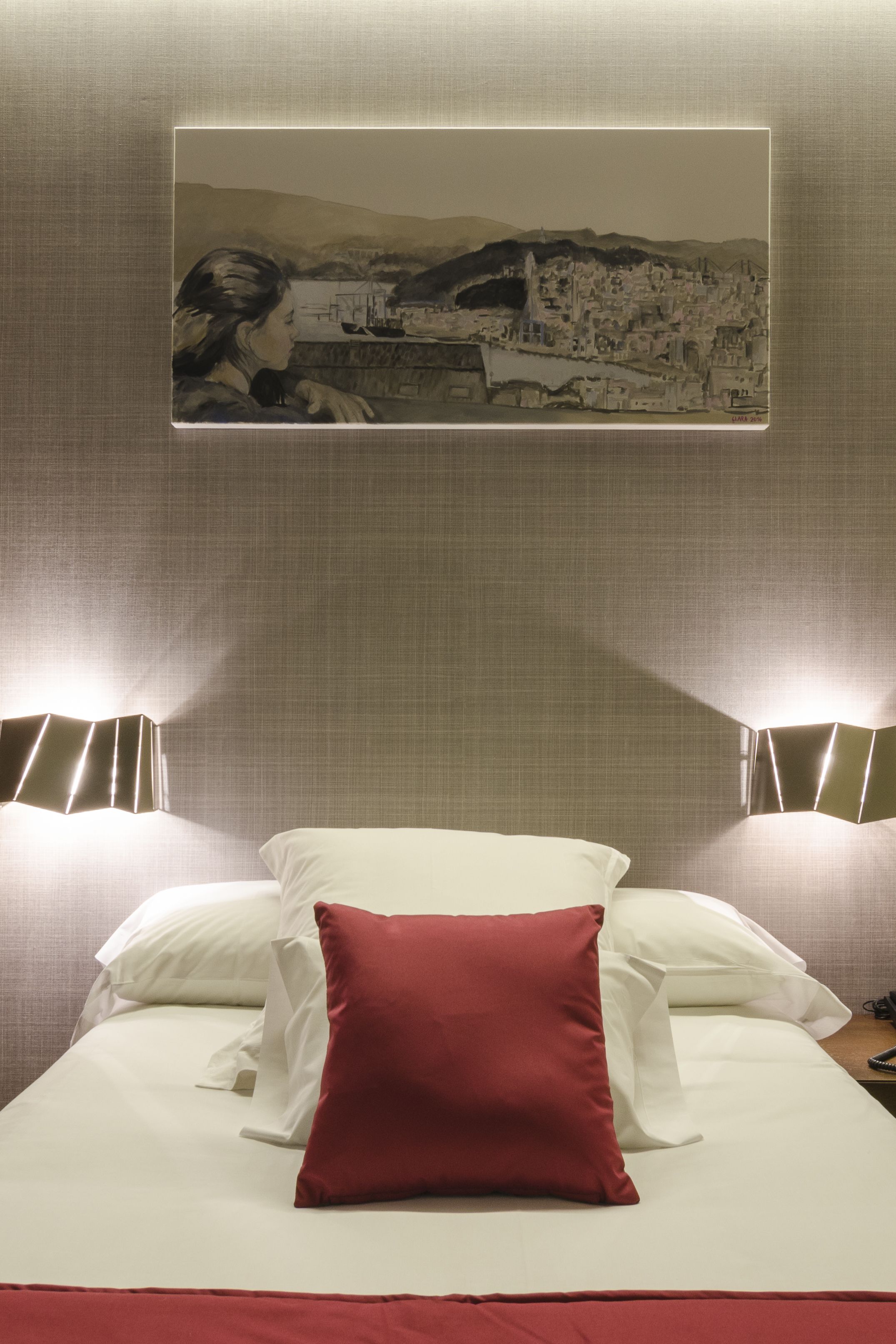 Una habitación de hotel con una cama, un almohadón rojo, dos lámparas de noche y un cuadro en la pared.