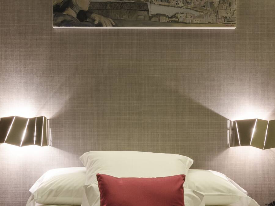 Una habitación de hotel con una cama, un almohadón rojo, dos lámparas de noche y un cuadro en la pared.