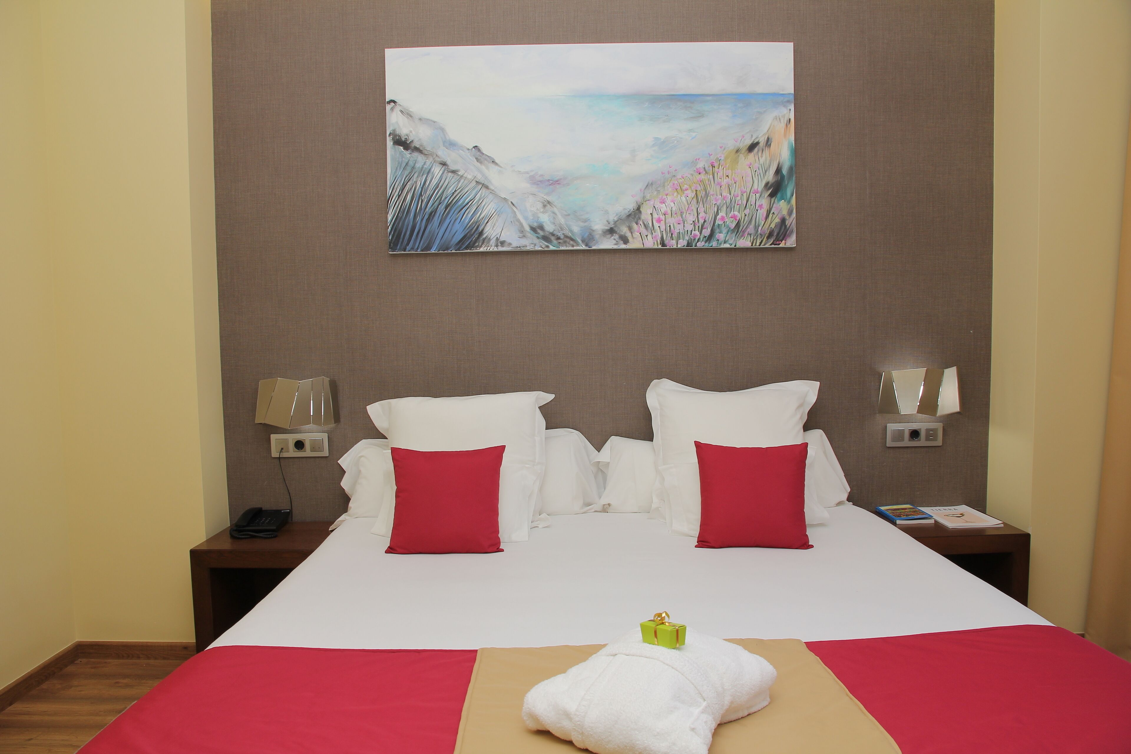 A hotel room with a large bed, two red pillows, a white blanket, a painting of a coastal scene, and a nightstand with a lamp and a phone.