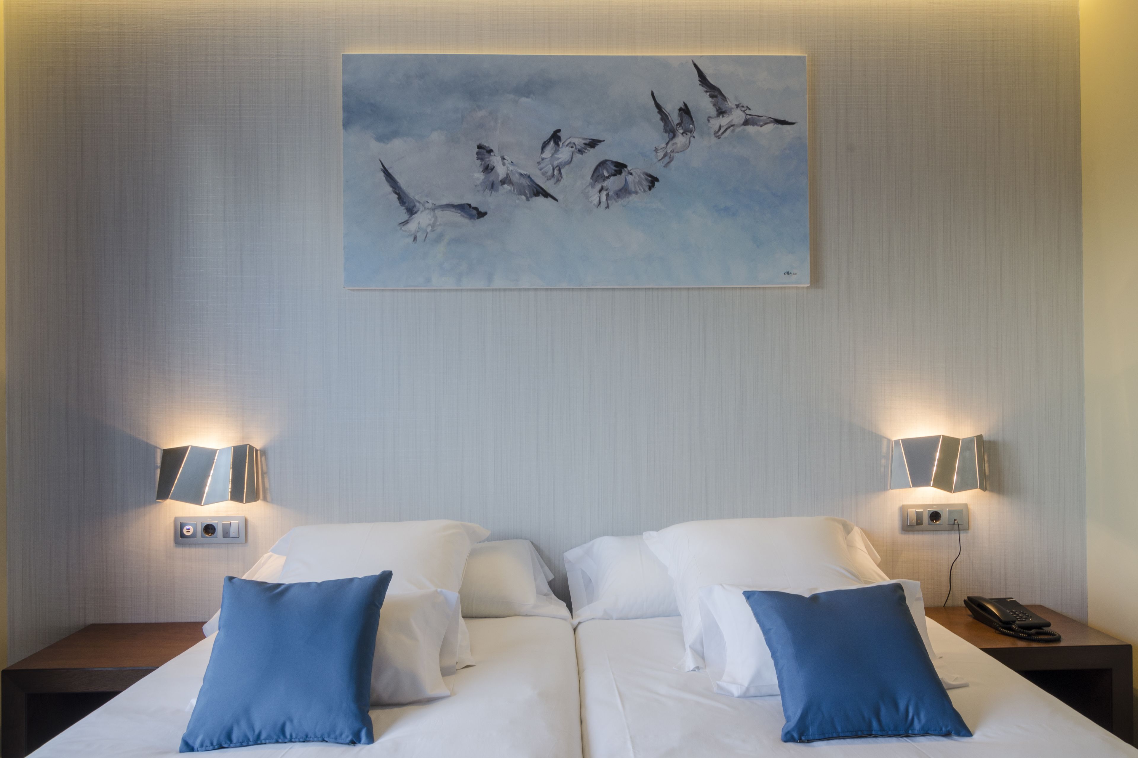 Habitación de hotel con dos camas, cada una con ropa blanca y almohadas azules, un cuadro de pájaros en la pared y lámparas de noche.