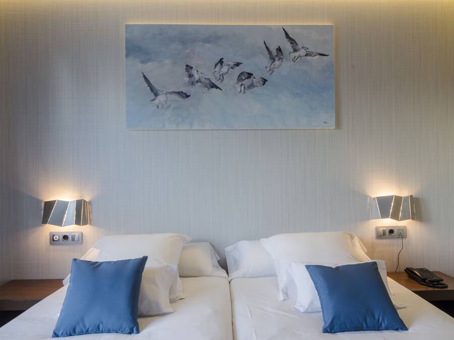 Habitación de hotel con dos camas, cada una con ropa blanca y almohadas azules, un cuadro de pájaros en la pared y lámparas de noche.